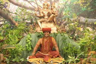 A seated figure in a red-orange turban, saffron-red robes, multiple rudraksha mala necklaces and orange armbands sits cross-legged on a tiger-skin mat in front of a green draped platform. Behind the figure a large golden statue depicts a seated deity with multiple arms and a companion figure, surrounded by lush artificial jungle foliage, branches and rocks. The person smiles broadly and rests a hand on the mat. Behind the figure, the painted backdrop shows a dense forest scene. Probable Swamiji cues: red turban, rudraksha malas and saffron-red attire, seated as a teacher on a tiger-skin seat.