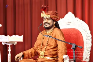 A smiling man in an orange-saffron silk kurta and a saffron turban with a red feather and gold jeweled brooch sits on an ornate white and red throne-style chair before red velvet drapes. He wears long gold-beaded and rudraksha-style necklaces, a gold ceremonial pendant, a gold waist belt and a gold earring, and is seated behind a microphone on a stand. A carved wooden lectern stands at the left edge. a person is seated in front of a microphone, speaking during a discourse. Watermarked archive photo.