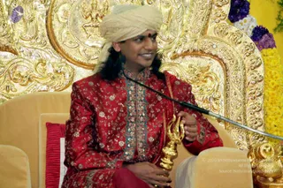 A man seated on a cushioned golden throne in front of an ornate gold-embossed backdrop with a flower garland of purple, white and yellow blooms at upper right. He wears a cream turban with a red bindi, and a red embroidered silk ensemble with gold and teal trim. He holds a long ornate gold staff or rod across his lap, smiling. A gold ornamental figure is partly visible at the lower right.