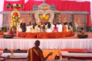 A photograph of a ceremony on a raised stage draped in red, orange and white cloth. At centre, a seated figure in a red and gold garment with a large garland and a crown-like headpiece sits before an ornate shrine with a grey carved temple facade and a golden kalasha finial. A large heart-shaped halo of coloured beads frames the central figure. Around the stage, rows of seated devotees in white and cream clothing, with some in yellow and pink, face the centre. A man in orange robes sits at the left beside a golden throne-like seat with a striped canopy and garlands. In the foreground, a shaved-headed priest in an orange-brown dhoti and shawl, with a sacred thread, holds a lit oil lamp aloft. Brass vessels stand on the stage edges.