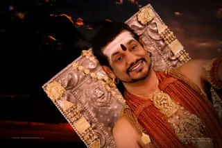 A close-up photograph of a smiling man with a white sandalwood-paste tripundra and a dark round bindi-like mark on his forehead, wearing layered coral-red beaded necklaces, a large gold pendant and a gold armband. Behind him are ornate silver-toned panels with embossed deity imagery and gold trim, against a dramatic dusk sky with orange clouds. The bare shoulder and rich adornments suggest a staged ceremonial portrait.