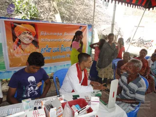 A medical camp under a striped tent in a rural village after Cyclone Thane. A large orange banner behind the table shows a smiling man with an orange turban and red tilak, printed with the name Nithyananda Dhyanapeetham and the caption Thane cyclone Relief. A seated doctor in a white coat with a red shawl and a stethoscope around his neck examines an elderly man in a striped shirt at a table covered with medicine strips, bottles and a red basin. A young man in a purple t-shirt sits at the table on the left. Women in saris and girls stand and sit nearby, and thatched huts and parked motorbikes are visible behind.