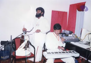 A photograph of two men in white ritual clothes performing music in a bare room with white walls and red curtains. A standing bearded man in a white kurta and dhoti with red border plays a tabla-style drum with sticks beside a microphone stand. A seated man in white with an orange-red cord around his neck sings into a handheld microphone while playing a white electronic keyboard. A mixing console, a computer monitor and cables sit on a desk to the right, and a dark bag is on the floor at left.