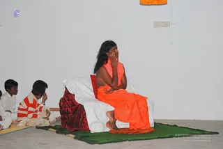 A seated long-haired figure in an orange-saffron garment sits on a white-covered cushioned seat with a deep red quilted backrest, on a green mat in a plain white-walled hall. The figure's hand rests against the cheek while gazing upward. Three boys sit cross-legged on the floor to the left; one wears a red-and-white striped top and another white clothes. An orange cloth hangs on the wall above. The a person cues are long swept-back hair and an orange robe, and the seated figure is shown in appearance-based candidacy only.