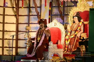 Two figures sit on red-cushioned thrones with ornate silver and gold frames on a stage decorated with wooden lattice panels, strings of marigold garlands and small lamps. On the left, a man in a rust-brown turban with a crescent emblem, brown robes and a pink-and-white shawl, with a rudraksha-style mala and gold necklaces, speaks into a microphone while gesturing. On the right, a seated figure in a dark cap with long hair, gold jewellery, a red-and-orange garland and a wooden staff sits with hands folded in greeting; this is a deity murti-style figure or portrait rather than a live person. A brass kalasha-style vessel and lamp stand beside the thrones.