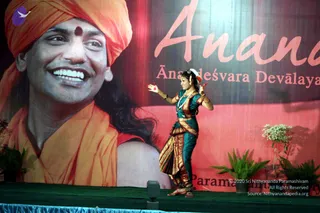 A female dancer in a teal bodice and gold-bordered red and gold skirt performs on a green-carpeted stage, arms extended with a red flower in her hand and a white flower in her hair. Behind her a large banner shows a smiling man in an orange turban and orange shawl, with a red bindi, labelled as Ananda Ānandeśvara Devālaya, with plants flanking the stage. a person depiction appears on the banner, not as a physical person, so the only physical person present is the dancer.