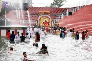 Outdoor water ceremony at a red-tiered amphitheatre-style pool. A seated figure in saffron-orange robes sits on an ornate golden throne under a decorated arch with floral garlands, while a fountain sprays water beside the steps. Numerous devotees stand waist-deep in the water, many women in white, red and green saris and men in white dhotis, some holding offerings. Children splash in the foreground. The setting resembles a Dasara celebration with a ritual bathing or abhishekam-style water rite.