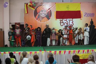 A stage performance by children in costume in front of a backdrop bearing a circular portrait of a smiling man in an orange-saffron turban and robe, with the year 2010 printed at upper right and a yellow-and-red flag hanging above a ring of marigold and green garlands. About a dozen children stand in a row on a green stage, dressed in regional costumes: one in green with a fan-like prop, one in orange with a bold patterned top, one in dark dress with a white shawl, others in white dhoti-style outfits, orange robes and a child holding a book. Some hold instruments such as a stringed instrument and a drum-like item. Audience members, including several adults and children, sit in the foreground. Balloons and a silhouette of a temple tower decorate the backdrop.