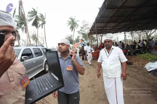 Outdoor relief work scene under palm trees on a dirt track. In the foreground a man in a white head cover holds up a dark laptop open toward the camera. Beside him a man in a grey-blue polo shirt and dark trousers, wearing a white head covering and a 'VOLUNTEER' badge, talks on a mobile phone while holding the laptop. On the right a man in a white shirt and white dhoti with a lanyard stands near a corrugated-roof shelter where people sit and gather produce. A silver SUV is parked to the left.
