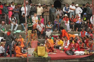 Outdoor ritual on stone steps at a riverside temple ghat, with a crowd of onlookers standing and seated on tiers above. In the centre, a woman in a peach-pink sari with flower garlands sits on a gold-framed seat, while saffron-robed priests in orange cloth sit around a red mat with brass pots, lamps and offerings. A cameraman on tripod films the scene at left. Many spectators wear red or orange shawls and ornate ceremonial dress. The setting appears to be a sacred bathing place with carved stone walls behind.