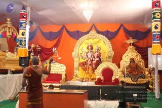 Photograph of a satsang stage under an orange and purple draped canopy, with a large gilded arch framing a deity murti pair seated on a red-cushioned throne, one figure with a crown and a long garland, the other with a decorated headdress. To the right, a gilded throne holds a small seated figure in orange robes with a rudraksha mala. In the foreground, a man with a shaved head wearing an ochre-brown dhoti and shawl raises one arm toward the altar beside a wooden lectern. Colourful striped pillars, a crystal chandelier, and monitors complete the scene.