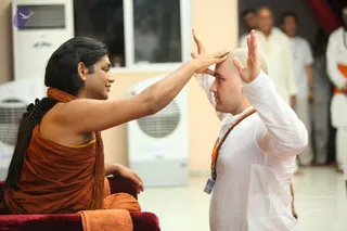 A seated man in saffron-orange robes with long dark hair, a rudraksha mala around his neck, sits on a red cushioned chair and extends his right hand toward the forehead of a bald man in a white kurta with an orange-beaded mala and an ID badge. The bald man holds his hands raised beside his head in a blessing posture. Behind them, several people in white and cream clothing stand near white air coolers in a bright room with a peach wall.