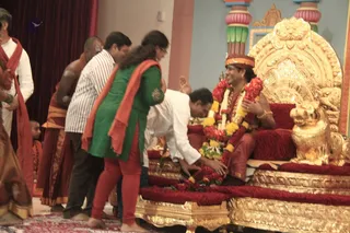 A photograph of a devotional reception in a temple hall. A man in a red-and-maroon dhoti and turban, wearing a large red and yellow marigold garland and gold bangles, sits on an ornate gold-and-red cushioned throne with a golden lion armrest. Several devotees stand and bend forward; one man in a white shirt and white dhoti leans down to touch the feet of the seated figure. A woman in a green kurta with orange shawl and a man in a striped shirt stand nearby with folded hands, and a priest in an orange-brown dhoti stands at left. Gold pillars with pink lotus capitals frame the background. Only the seated figure's appearance is consistent with the page's named teacher, so the identity remains uncertain.