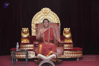 A man with short hair and a red bindi sits on a brown leather throne with an ornate gold arched backrest flanked by two gold lion-shaped armrests on a raised gold-trimmed platform. He wears a red saffron-toned robe with gold borders, a long beaded garland of gold-coloured beads, and a small microphone on his face, gesturing with both hands while speaking. A bottle of water sits on a small black side table to the left, and a wooden footstool rests in front. Dark red velvet drapes form the backdrop and a red carpet covers the stage.