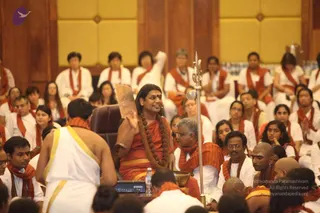 A seated speaker in a saffron-orange robe with a long rudraksha mala and a golden necklace sits in a brown leather chair on a raised platform, raising one hand while addressing the hall. Before him a man in white dhoti with gold border and orange shawl stands with his back to the camera. A brass trident-shaped staff and a brass bowl sit beside the platform. Rows of seated devotees in white clothes with orange shawls fill the wooden-panelled hall behind, some with hands together.