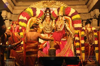 A photograph of a temple shrine with a golden deity murti pair, Shiva and Parvati, adorned with multicolour flower garlands in yellow, green, purple and red and heavy gold jewellery. The deity on the left holds a trident. Two priests in saffron-orange dhotis, one bare-chested with a rudraksha mala, stand at the left, one holding a lamp; a priest in a red dhoti and shawl with a sacred thread faces the shrine in the centre, performing worship. Golden wall carvings frame the altar, and a standing golden statue on the right in a dark shirt is garlanded.
