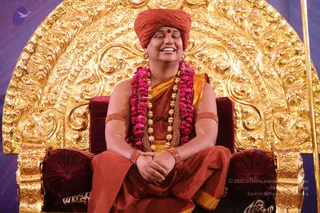 A seated figure on an ornate gold throne with a large embossed gold back, wearing a rust-orange turban with a red bindi, a pink flower garland, a long gold-bead mala, orange-and-gold robes, and gold bangles on the arms. He sits cross-legged with hands folded in his lap, eyes closed and smiling broadly. Red velvet cushions line the throne arms. Setting appears to be a stage during a darshan or satsang. One physical person is visible.