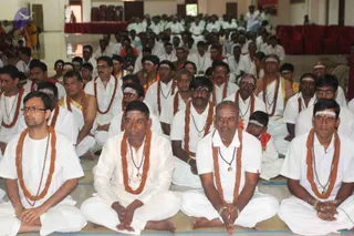 Photograph of a large seated congregation in a hall with white floor mats. Front-row men sit cross-legged in white dhotis and white shirts, wearing orange-brown rudraksha-style prayer garlands and beaded necklaces, with white tilak lines on their foreheads. Many in the rows behind wear similar white attire and garlands; a child sits among them. Rows of white-clad devotees fill the background toward a stage with red draped seating. No individual is identified as a person; the scene shows devotees attending a satsang-style gathering.