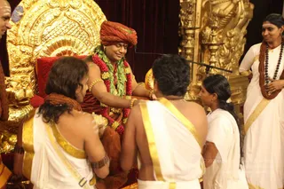 A photograph of a ritual scene on a gold-ornate throne. A figure seated in the centre wears a red-orange turban, a wrap of the same colour, and multiple garlands of red flowers and green leaves, with gold armlets and bangles. Two devotees in white dresses with gold borders sit or kneel before him, one holding a tray of offerings. A woman in a white sari with gold border and a rudraksha mala stands at right beside a gold ornate pillar, and a girl in white stands at centre-right. A man in the top-left corner wears a white garment. The seated figure has a crown-like turban and bright golden throne; the photo shows a Deepavali and Kali puja-related event. a person seated on a throne with long swept-back hair and a gold-and-red adornments, receiving offerings.