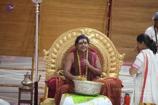 A photograph of a man seated on an ornate gold throne with a carved, arched back, wearing a maroon robe and a brown shawl draped over his lap, with a red tilak on his forehead, a rudraksha mala and a gold-toned garland around his neck. He holds a small stick in one hand over a large silver bowl filled with yellow flowers or offerings, while smiling. To the right, a woman in a white sari with a red-and-pink border stands with palms pressed together in namaste. Behind them is a bamboo-screen backdrop, a tall silver lamp stand, and a silver vessel on a small wooden table. Several people are partly visible at the edges. The scene suggests a puja or ritual offering.