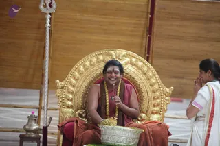 A seated man on an ornate gold throne smiles broadly and holds a small lit item between his fingers above a silver bowl. He wears a maroon sleeveless robe, a brown shawl draped at the waist, a bright red bindi and a marigold garland with a rudraksha-style mala. Beside him stands a silver trident and a brass vessel with a bottle on a small table. A woman on the right, in a white sari with a red border, stands with palms pressed together in namaste. Wooden bamboo screens form the backdrop. The scene appears to be a puja or darshan. a person is seated on the throne with long swept-back hair, a maroon robe and a rudraksha mala.
