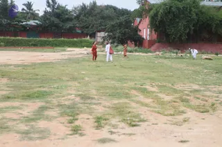 A photograph of a large open grassy and dirt field on an ashram campus, with patches of worn grass and scattered stones. Four or five people are visible: a woman in orange clothes stands at left in conversation with a woman in white garments carrying a red bag, while a younger person in orange walks nearby and two people in white clothes bend down to work near a red-walled building at right. Dense green trees and a red building with white-framed windows fill the background.