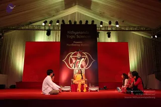 A stage session under a white tent with a red stage floor and red backdrop panels. A woman in a yellow-orange sari with a gold necklace sits on an ornate silver throne in the centre. Beside her stands a large banner reading Nithyananda Yogic Sciences Presents Manifesting Shaktis, showing a bearded man with a red turban, white garland and a raised hand over a golden yantra-like emblem. Two people in white shirts sit cross-legged on the left, and three women in red and dark clothing sit on the right, facing the throne. Stage lights hang from a truss above.
