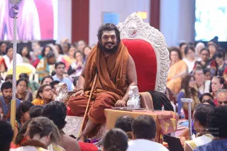 A photograph of a bearded man with long dark hair seated on an ornate silver-and-red throne with carved lion heads, smiling and holding a wooden staff. He wears a saffron-orange robe and a small microphone headset. Before him, a large audience fills rows of seats, including many women in orange, yellow and saffron saris and men in casual clothes. Large screens behind show pink and blue imagery. Silver ceremonial staffs stand at the left and right edges of the stage, and a low table draped in gold and red cloth sits in front of the throne.