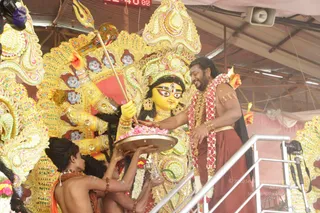 A photograph of a large, ornately decorated golden Durga murti with a yellow face, many arms and a crown, set in a temporary open-sided pandal under a corrugated roof. A long-haired man with a long beard wears a red-maroon dhoti, a red and white flower garland, gold armbands and bangles, and holds a brass plate of pink flowers toward the deity while a trident-bearing arm of the murti is nearby. Two shirtless attendants in orange cloth and rudraksha malas stand below the platform, one holding the plate and another holding a small brass vessel. Pink and white flower offerings are visible at the base, and a loudspeaker and camera are on the right. Two people are visible in the scene.