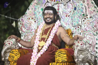 A seated man in dark red-brown robes smiles broadly on an ornate silver throne encrusted with red, green and blue gemstones, with elephant-head armrests draped in tiger-striped cushions. He wears layered gold necklaces, a pink and white flower garland, gold armlets and bangles, and a white-painted forehead with a central mark. Behind him is a blurred dark background with a thin cable. Swamiji (probable): the seated man has long swept-back hair, a beard, ornate gold jewellery and saffron-toned robes on a jewelled throne, a ceremonial presentation rather than an everyday pose.