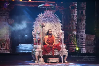 A seated man in an orange-red saffron robe and a long rudraksha mala sits on an ornate silver throne with lion-headed armrests and a silver trident on a stand beside him, under a silver canopy. Stone temple columns carved with figures frame a stage lit with blue and warm light, with smoke drifting at left. A person with long hair and an ornate silver throne is the central figure, hands folded in his lap. One person is visible.