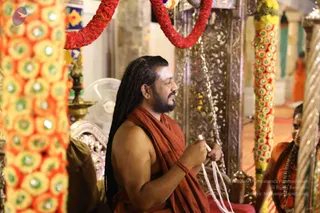 A man with long dreadlocked hair and a trimmed beard sits on a red-cushioned seat, bare-chested except for a rust-orange shawl draped over his shoulders, holding a white cord or thread in his hands while looking to his right. Behind him, an ornate silver-toned throne-like backrest is framed by garlands of yellow and orange marigolds and red flower garlands strung overhead. A brass oil lamp stands at left, and a partially visible woman in orange clothing with a rudraksha-style necklace sits at the right edge. A man in orange robes stands in the background. a person: the seated man has long dreadlocks, an orange shawl and a ceremonial setting, consistent with the page's a person reference.