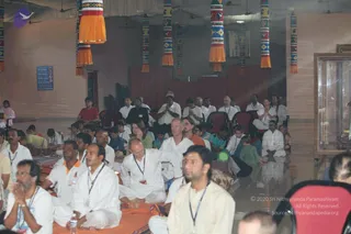A large indoor satsang hall filled with seated devotees in white clothes, many wearing blue lanyards, sitting on orange and mustard cushions on the floor. Rows of red chairs are at the back and several people hold their palms together in anjali. Colourful woven hanging banners with orange tassels decorate the ceiling. A small display case is on the right wall. The scene looks like a satsang or meditation session. No clearly identifiable a person figure is visible in this view, so the role of the speaker is not shown.