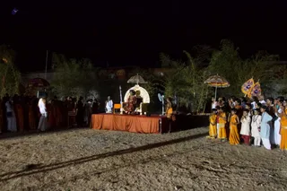 Night outdoor photo of a ritual gathering on a dirt ground beside bamboo and a low wall. A raised platform draped in red-orange cloth holds a seated figure in red-orange robes before an ornate white arched backdrop with a white parasol above. Men in white dhotis stand near the platform, and a woman in an orange sari stands at its side. On the right, a line of children and young people in orange and yellow clothing face the platform, some holding small white and orange parasols. Two decorated flags on poles are visible at right. Several people stand at left in orange and white attire. No identity is claimed for the seated figure.