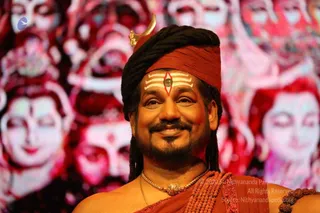 Close-up photo of a smiling man with long dark dreadlocks wrapped under a rust-red headband topped by a small golden crescent ornament. He has white horizontal tripundra lines with a red-and-black central mark on his forehead, a dark beard, and a gold pendant necklace over a rust-orange cloth draped over his shoulders with rudraksha beads visible. Behind him is a large backdrop of repeated pink-red stylised faces. a person appearance cues are present (long matted hair, crown-like ornament, saffron-orange draped cloth, ceremonial gold necklace), so this is a probable in-person candidate.