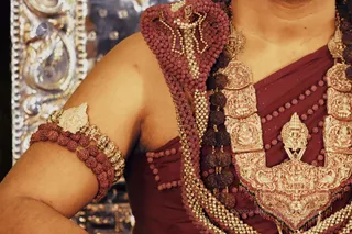 Close-up photograph of a bare-shouldered figure from the chest to upper arm, wearing a deep maroon garment. Heavy gold ceremonial jewellery covers the chest: a large embossed gold necklace with figurative panels and a central pendant, layered with dark and red rudraksha malas and a red beaded cord. A gold armlet with leaf motifs sits on the upper arm, with rudraksha beads on the wrist. Behind, an ornate silver embossed surface reflects light. A small logo and a faint copyright line appear at the lower edge. The face is not visible, so the figure is identified only by these cues.