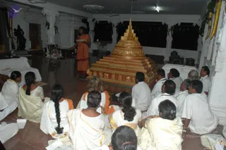 Indoor temple hall with white marble walls and dark windows at night. A golden tiered pyramid-shaped structure. A standing woman in an orange saffron robe reads from a sheet of paper beside it. Around the structure, a group of devotees sit on the polished floor in white and cream ritual clothes, some with gold-bordered shawls, many with hair in braids, some holding papers. A dark deity murti stands on the left near a white pillar, and a smaller figure sits on a pedestal at right. Garlands hang near the upper right windows.