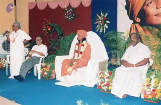Stage event with a white-draped chair at centre. A seated figure wears an orange turban, orange robe with gold trim, and a garland of white and red flowers over a rudraksha-style mala; he is seated, hands folded. A man in white dhoti and shirt stands at left speaking into a microphone, holding a lapel badge. Two other men in white shirts sit beside, one in grey trousers and glasses, one at right in white dhoti. Blue carpeted stage with pink and maroon drapes, palm plants, flower arrangements, and a large portrait of a turbaned face in orange at upper right. a person (in_person).