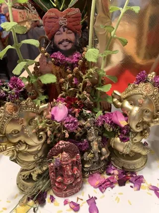 A cut-out cardboard figure of a smiling man in an orange-red turban with a gold emblem and a rudraksha mala stands behind a shrine arrangement. Two gold-finished seated Ganesha figurines flank a small red Ganesha relief and a dark bronze Ganesha statue. Purple chrysanthemum flowers, pink petals, green leaves and scattered yellow rice cover the white table. No people are physically present.