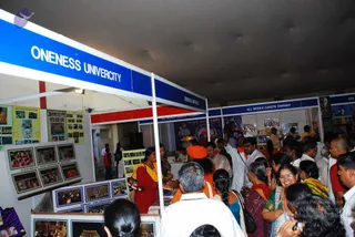 A busy photograph of an exhibition hall at a spiritual service fair. A blue-and-white booth header reads ONENESS UNIVERCITY, with a red-and-white booth beside it labelled ALL WORLD GAYATRI PARIWAR. A woman in a red salwar kameez stands at the front of the Oneness booth, and a man in white shirt stands among visitors. A man in an orange turban and saffron clothing stands near the centre of the crowd, and a few people in white and orange clothes are visible. Display panels show flood relief and free clothing photographs, homa photographs and posters. Many visitors in sarees, shirts and kurtas crowd the aisle and look at the displays.