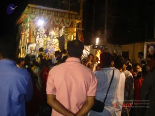 Night photograph of a procession at a temple-style ratha (chariot) with ornate gold-painted carved pillars, a golden canopy with red and white draped fabric, and deity murti decorated with white and yellow flower garlands. A bright floodlight and an open flame of a torch or lamp are visible. A crowd of devotees stands close around the ratha; a man in a pink polo shirt faces away in the foreground, another man in a light blue striped shirt wears a black shoulder bag, and women in red and other saris are visible on the right. A large printed portrait banner appears at the right edge. Scaffolding and a tented enclosure sit in the background.
