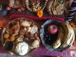 Overhead photo of a food offering laid before a deity murti decorated with gold and jewelled garlands, a gold crown-like ornament, and a rudraksha bead garland. On a silver thali are fried snacks, round sweets, cooked white rice, and a steel cup, with a cup of white paste. Beside it is a dark leaf bowl with a red apple, dark grapes and ripe bananas. Marigold flowers and a red cloth are visible. No people are visible.