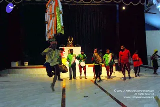 A photograph of a stage performance in an indoor hall with dark drapes and a white-tiled floor. In the foreground a man in a yellow-and-white patterned shirt and a red head cloth leaps mid-air while holding a drum. Behind him, six men stand in a line; several wear bright green shirts with red-and-maroon dhoti-style lower garments and play hand drums, while a man in a red shirt and dark dhoti stands at the right and another in red and saffron stands at the far right edge. A small deity murti or statue garlanded with a yellow-orange garland sits on a white-draped platform, with a hanging orange, white and green floral garland above it. Potted plants flank the stage.