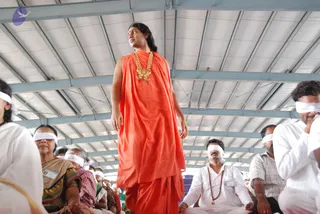 A standing man in a flowing saffron-orange robe and a gold pendant necklace strung with beads stands above a seated crowd inside a metal-framed hall with a white corrugated roof. Many seated people wear white head bands covering their eyes, including a woman in a patterned sari and men in white shirts, one wearing a red beaded mala. A man in a white kurta sits at right with hands together. Blue steel beams cross the ceiling and a purple logo sits at upper left. The standing figure shows long swept-back dark hair and distinctive orange-and-gold attire, but the appearance is not conclusive enough to confirm identity, so the figure is described by role as a robed presenter.