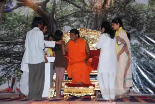 A seated man in an orange-saffron robe sits on an ornate gold throne with red cushions in front of a painted backdrop of a large banyan tree. Several people in white and cream clothing stand around him on a stage. A man in a white shirt and grey trousers reaches toward the seated man's shoulder, a young girl in a brown top and striped pants stands close by, and two women, one in white churidar and one in a cream sari with a yellow shawl, stand at the sides. A white pedestal is visible on the left and a small child in yellow appears behind the girl. The seated man has short dark hair and no visible long hair, crown, or rudraksha mala, so his identity is uncertain.