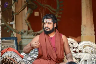 A bearded man with long dark hair and white forehead markings with a central dot sits on an ornate silver chair in a courtyard, wearing a maroon-brown shawl over a dark red sleeveless top. He gestures with his right hand while speaking. Behind him stands a bronze statue of a dancing deity with raised arms and a flame-like arch, set against a red wall and a stone pillar. A leopard-print cloth lies on the chair's left side. A small microphone is clipped to his top. Appearance-based label: a person, seated and speaking as a teacher in the courtyard.
