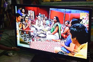 A flat-screen monitor on a stand displays a live video feed of a ritual gathering in a red-walled tent. Seated on a red and white patterned mat, a man in a green and white garment sits centrally facing a group of devotees, several with hands in anjali; a woman in red and a woman in striped sari with a bindi are among them. A shaved-headed man in white kneels in folded-hands greeting. Behind them stand additional participants and a framed deity murti on a shelf. In the foreground, the back of a person in a yellow top watches the screen, and an overlay menu with icons is visible on the display. Ritual setting shows devotees gathered around a central seated figure.