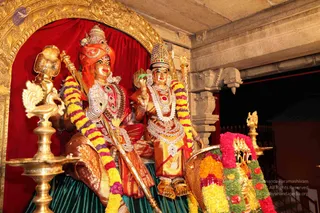 A photograph of two decorated murti figures seated on a green-draped platform within a golden-framed niche with a red velvet backdrop. The figure on the left wears an orange turban, a red and white ornate garment, a large beaded necklace, and holds a tall staff. The figure on the right wears a gold and red crown, layered white and gold garlands, and holds a small ceremonial object. Both are adorned with flower garlands in yellow, purple, pink and green. Brass oil lamps stand on either side, and a flower-decorated palanquin or chariot section appears at right. These are statues, not physical people.