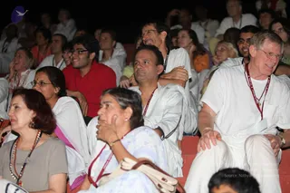 A seated audience at an indoor satsang, with rows of devotees in white clothing and red lanyards around their necks. In the foreground, a man in a white shirt and white trousers sits at right with a smile, while a woman in a light blue patterned salwar sits beside a woman in a grey top wearing a rudraksha-style mala. Behind them, a young man in a red shirt and glasses watches attentively, and a man in glasses laughs while embracing a companion in white. The stage and the speaker are not visible; the attendees face forward toward the event. Dim warm lighting and red-cushioned tiered seating surround the crowd.