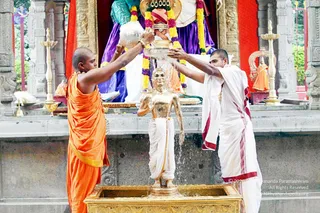 Temple abhishekam scene: two shaved-headed priests pour water over a golden murti of a standing sage figure on a gold-trimmed pedestal. The left priest wears a saffron/orange robe and holds a silver vessel; the right priest wears a white dhoti and shawl with maroon borders, holding a silver bowl with an upward gesture. Behind them, a deity murti draped in purple and white with garlands of yellow and magenta flowers and a red-and-gold headdress, flanked by brass lamps and red drapery. No identifiable a person figure is visible.